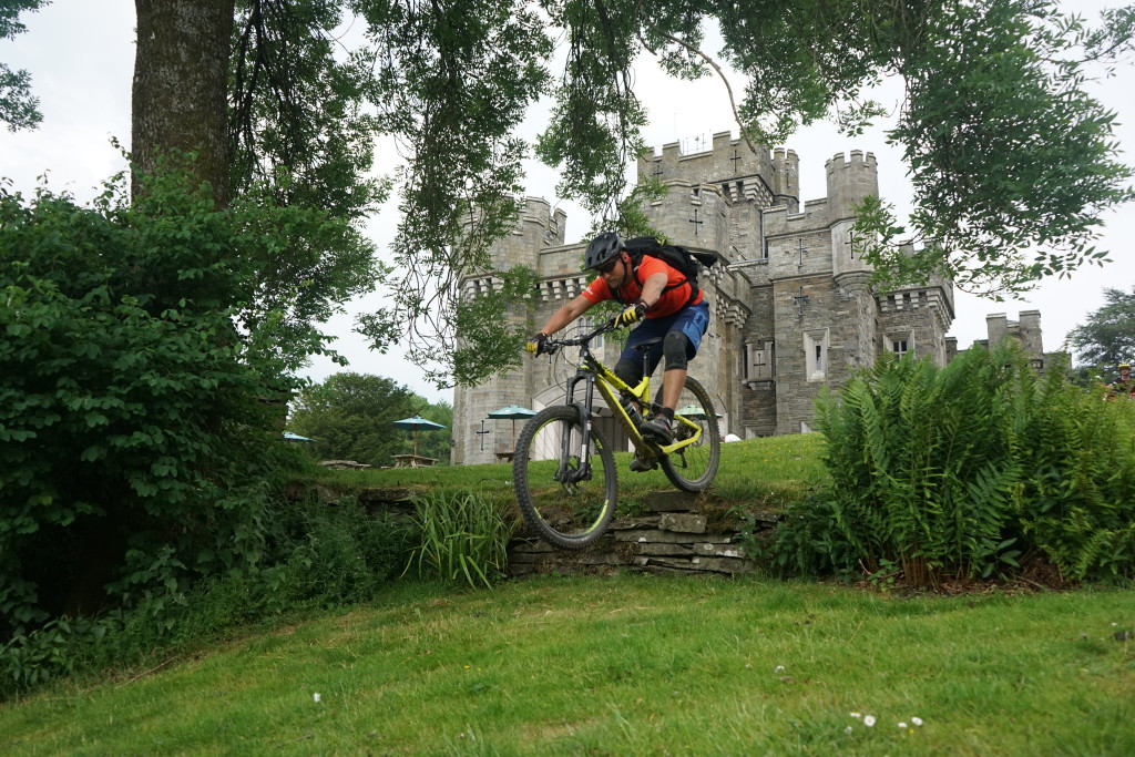 Rik Harms dropt zijn mountainbike van een kleine stenen muur op het gras. Op de achtergrond staat een mooi Engels kasteel in Lake District